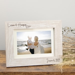 May include: A light-colored wood picture frame with a white mat, featuring the engraved text "Lucas & Harper" with a small leaf illustration at the top left and the date "June 6, 2025" engraved at the bottom right corner of the frame.