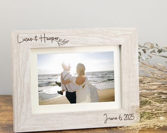May include: A light-coloured wood picture frame with a white mat, featuring the engraved text "Lucas & Harper" with a small leaf illustration at the top left and the date "June 6, 2025" engraved at the bottom right corner of the frame.