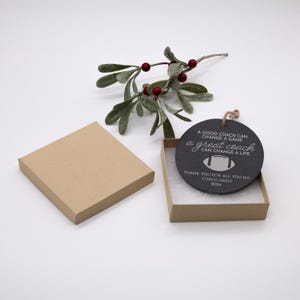 May include: A black round ornament with a white inscription that reads "A good coach can change a game, a great coach can change a life". The ornament also includes a white football graphic and the text "Thank you for all you do, Coach Gregi 2024". The ornament is in a brown box with white filling.