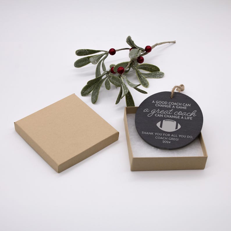 May include: A black round ornament with a white inscription that reads "A good coach can change a game, a great coach can change a life". The ornament also includes a white football graphic and the text "Thank you for all you do, Coach Gregi 2024". The ornament is in a brown box with white filling.