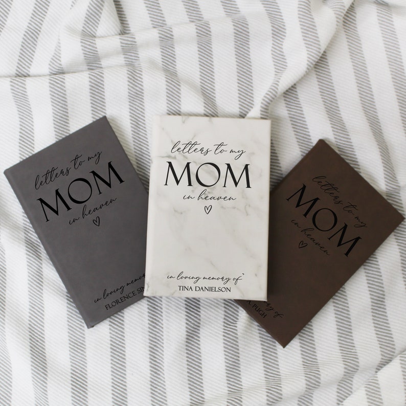 May include: Three grey, brown and white journals with marble covers. Each journal has the text "letters to my MOM in heaven" with a heart symbol. The white journal has the text "in loving memory of TINA DANIELSON".