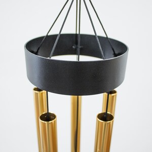 May include: A black metal wind chime with gold metal tubes hanging from it. The wind chime has a round top and is suspended from a black string.