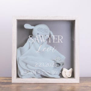 Peut inclure: Une boîte à ombre en bois blanc avec une couverture de bébé bleu clair, un jouet en peluche en forme d'agneau, une sucette et le texte "Sawyer Levi 2.23.2025 1:48 AM 7 lbs 11oz".