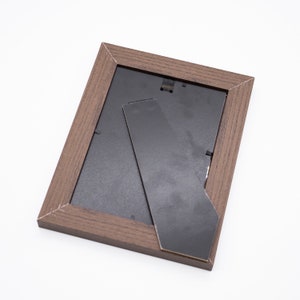 May include: A brown wooden picture frame with a black mat and a black easel stand on the back.