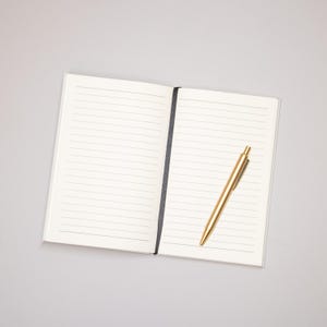 May include: An open notebook with lined pages and a gold pen lying on the right page.
