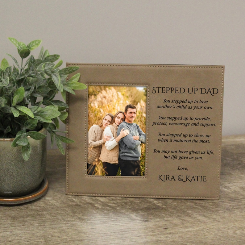 Peut inclure: Un cadre photo en cuir marron avec une photo de trois personnes. Le cadre porte le texte "Stepped Up Dad" et un message sur l'amour et le soutien d'un beau-p&egrave;re. Le message se termine par "Love, Kira & Katie".