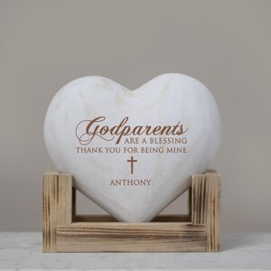May include: White heart-shaped stone with the text "Godparents are a blessing thank you for being mine." and a cross with the name "Anthony" below it. The stone is on a wooden stand.