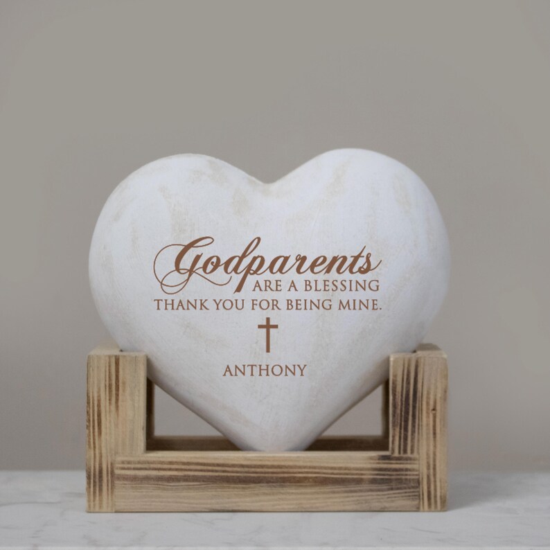 May include: White heart-shaped stone with the text "Godparents are a blessing thank you for being mine." and a cross with the name "Anthony" below it. The stone is on a wooden stand.