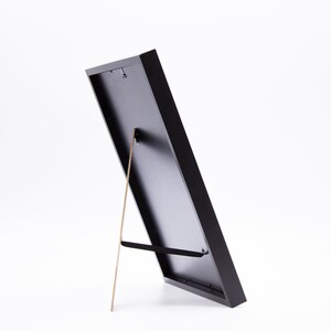 May include: A black picture frame with a built-in easel stand. The frame is made of wood and has a matte finish.