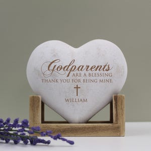 May include: White heart-shaped stone with the text "Godparents are a blessing thank you for being mine." and a small cross with the name "William" below it. The stone is on a wooden stand.