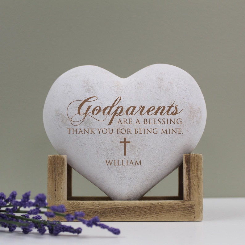 May include: White heart-shaped stone with the text "Godparents are a blessing thank you for being mine." and a small cross with the name "William" below it. The stone is on a wooden stand.