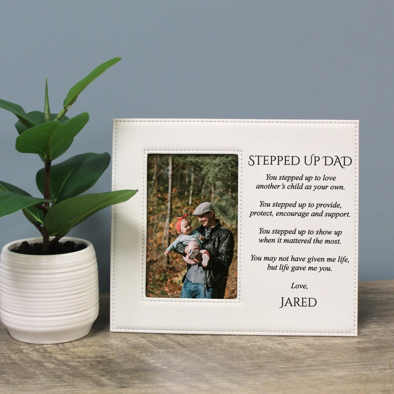 Peut inclure: Cadre photo blanc avec une bordure cousue et le texte "Stepped Up Dad" avec un message sur une figure paternelle. Le cadre comprend une photo d'un homme tenant un jeune enfant.