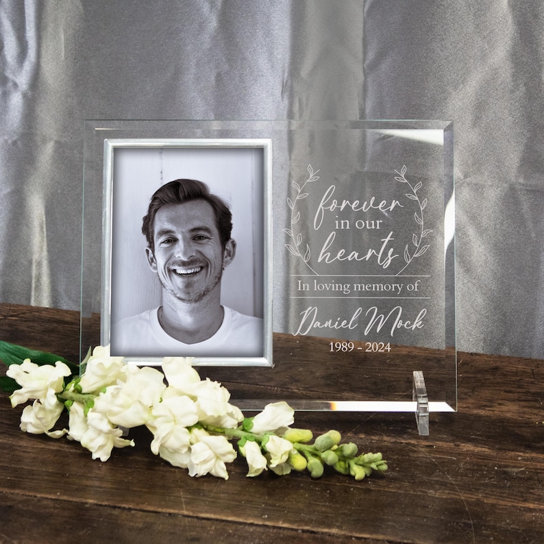 K&ouml;nnte beinhalten: Ein klarer Glas-Bilderrahmen mit silbernem Rand. Der Rahmen hat eine gravierte Inschrift mit dem Text "forever in our hearts" und "In loving memory of Daniel Mock 1989-2024".