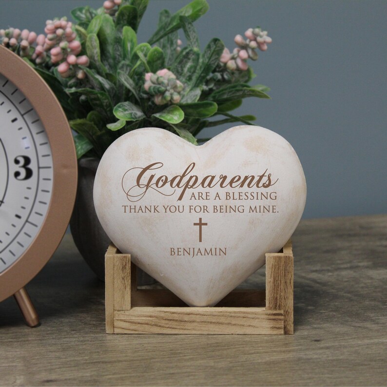 May include: White heart-shaped stone with the text "Godparents are a blessing thank you for being mine." and a small cross. The name "Benjamin" is written below the cross. The stone is sitting on a wooden stand.