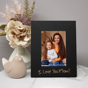May include: A black picture frame with a photo of a mother and child. The frame has the handwritten text "I Love You, Mom!" in gold. A decorative bird and floral arrangement are in the background.