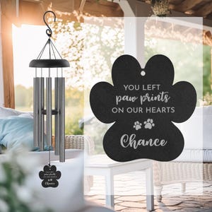 May include: A black wind chime with silver-colored tubes and a paw-shaped pendant. The pendant reads "YOU LEFT paw prints ON OUR HEARTS Chance" in white script. The wind chime hangs from a black hook.