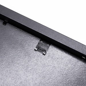 May include: Close-up of a black picture frame with a metal hanging clip. The frame has a textured surface.