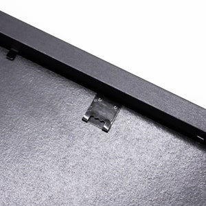 May include: Close-up of a black picture frame with a metal hanging clip. The frame has a textured surface.