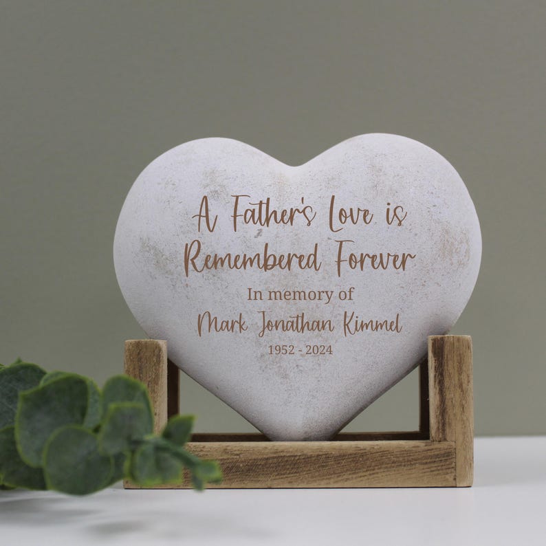 以下が含まれることがあります： 「A Father's Love is Remembered Forever」と「In memory of Mark Jonathan Kimmel 1952 - 2024」の刻印がされた白いハート型の石。石は木製の台座の上に置かれています。
