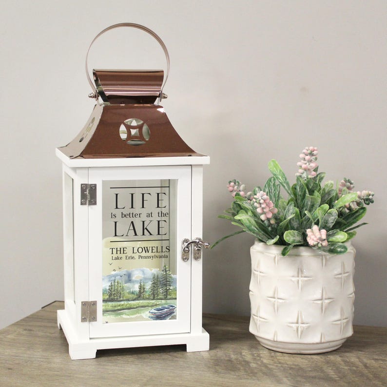 May include: White lantern with a copper top and a glass window. The glass window has a watercolor image of a lake scene and the text "Life is better at the lake. The Lowells. Lake Erie, Pennsylvania."