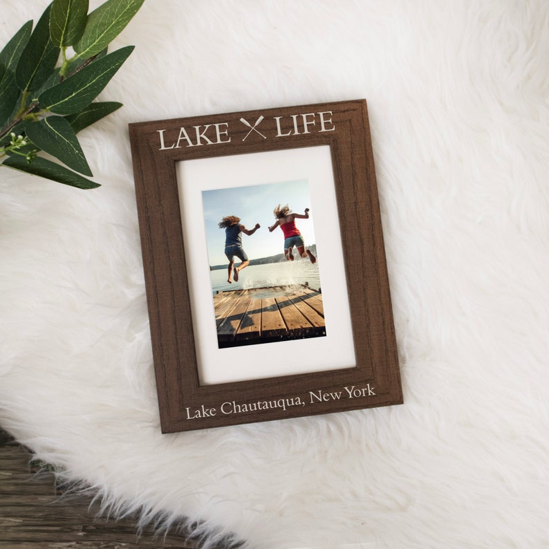 May include: A brown wooden picture frame with the text "LAKE LIFE" and two crossed oars above the text. The frame has a white mat and a photo of two people jumping off a dock into a lake. The text "Lake Chautauqua, New York" is printed below the photo.