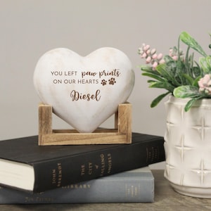 May include: A heart-shaped memorial stone with the text "YOU LEFT paw prints ON OUR HEARTS Diesel" and paw print graphics. The stone is supported by a wooden stand and rests on top of books. A vase of flowers is in the background.