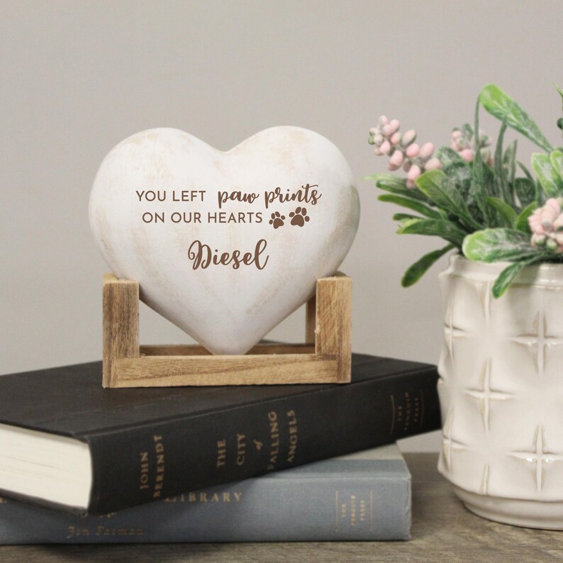 May include: A heart-shaped memorial stone with the text "YOU LEFT paw prints ON OUR HEARTS Diesel" and paw print graphics. The stone is supported by a wooden stand and rests on top of books. A vase of flowers is in the background.