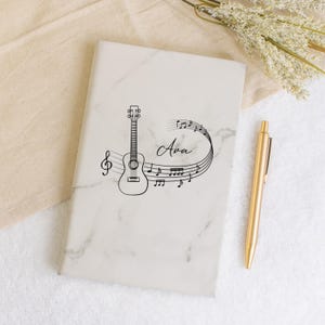 May include: A white marble notebook with a black ukulele and musical notes design. The name "Ava" is written in cursive on the notebook.