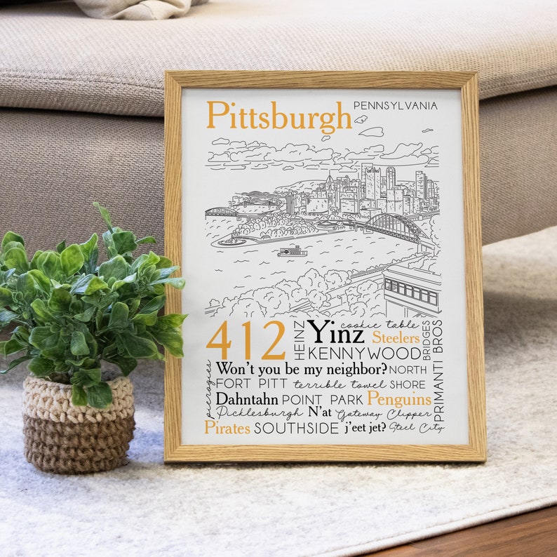 May include: A framed print with a black and white illustration of the Pittsburgh skyline. The print features the text "Pittsburgh, Pennsylvania" and a list of words and phrases associated with the city, including "412", "Yinz", "Steelers", "Kennywood", "Won't you be my neighbor?", "North Shore", "Fort Pitt", "terrible towel", "Dahntahn", "Point Park", "Penguins", "Picklesburgh", "N'at", "Gateway Clipper", "Pirates", "Southside", "j'eet jet?", "Steel City", "Bridges", and "Primanti Bros."