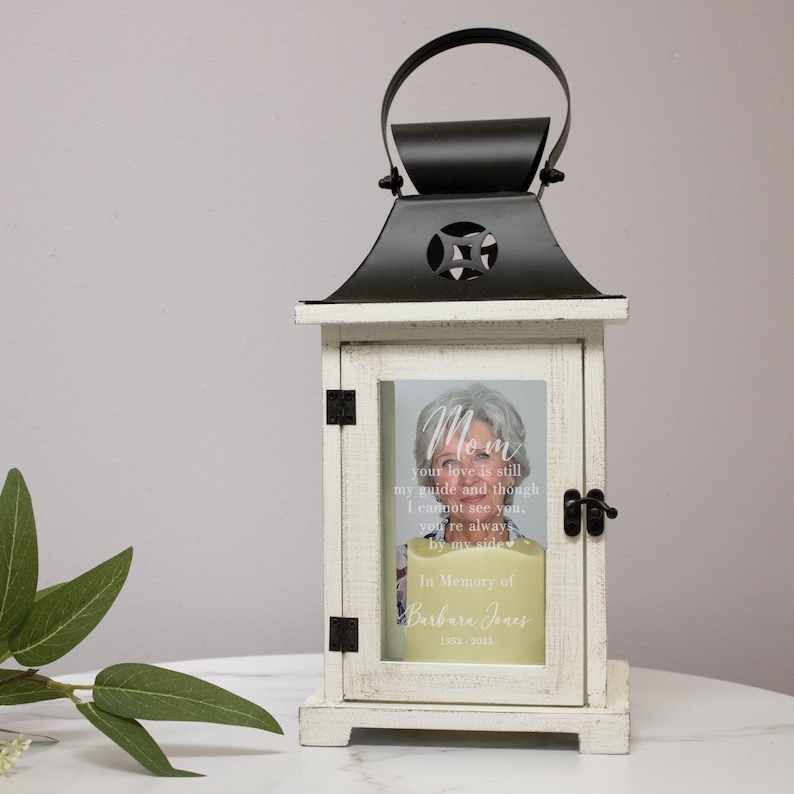 May include: White wooden lantern with a black metal top and a glass panel. The glass panel has a photo of a woman and a white inscription that reads "Mom your love is still my guide and though I cannot see you, you're always by my side. In Memory of Barbara Jones 1952-2015."