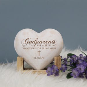May include: White heart-shaped stone with the text "Godparents are a blessing thank you for being mine." and a cross with the name "Teagan" below it. The stone is on a wooden stand.