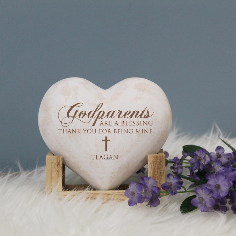 May include: White heart-shaped stone with the text "Godparents are a blessing thank you for being mine." and a cross with the name "Teagan" below it. The stone is on a wooden stand.