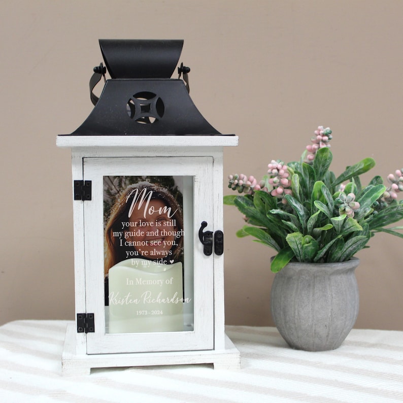 May include: White lantern with a black top and a glass window. The window has a photo of a woman and a message that reads "Mom your love is still my guide and though I cannot see you, you're always by my side. In Memory of Kristen Richardson 1973-2034".