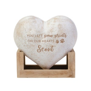May include: A heart-shaped memorial stone with the inscription "YOU LEFT paw prints ON OUR HEARTS Scout." The stone is off-white with a distressed finish and rests in a light brown wooden stand. Paw prints are also included in the inscription.