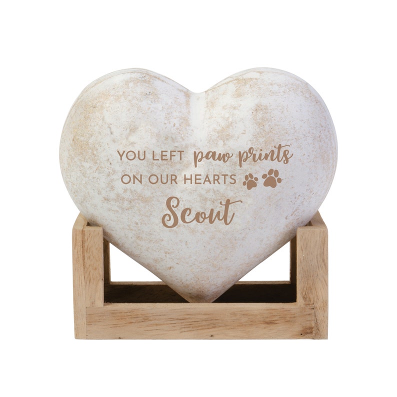 May include: A heart-shaped memorial stone with the inscription "YOU LEFT paw prints ON OUR HEARTS Scout." The stone is off-white with a distressed finish and rests in a light brown wooden stand. Paw prints are also included in the inscription.