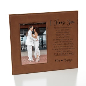 May include: A brown leatherette photo frame with a stitched border. The frame holds a photo of two people in white clothing embracing. The text "I Choose You" is engraved on the frame with a message about choosing someone over and over again. The names "Nia" and "Amaya" are also engraved on the frame.