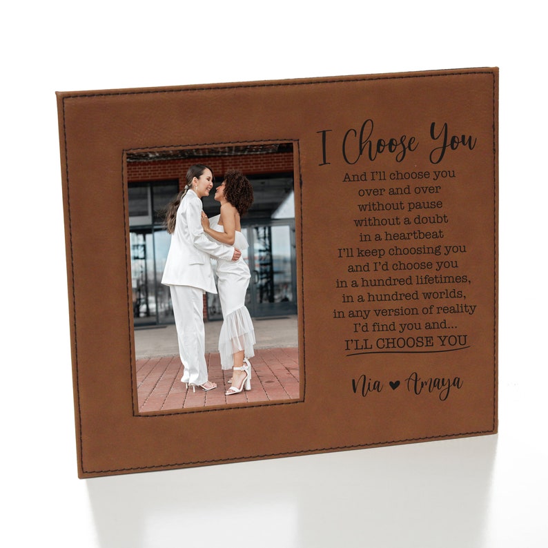 May include: A brown leatherette photo frame with a stitched border. The frame holds a photo of two people in white clothing embracing. The text "I Choose You" is engraved on the frame with a message about choosing someone over and over again. The names "Nia" and "Amaya" are also engraved on the frame.