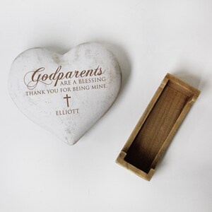 May include: White heart-shaped stone with the text "Godparents are a blessing. Thank you for being mine." and a cross. The name "Elliott" is written below the cross. The stone is sitting on a white surface.
