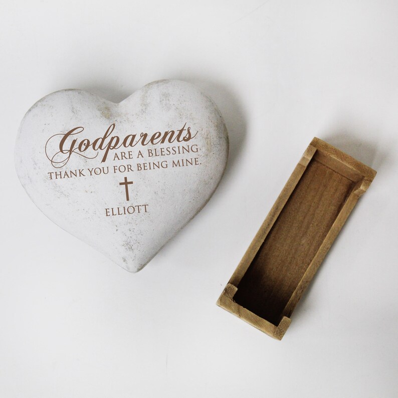 May include: White heart-shaped stone with the text "Godparents are a blessing. Thank you for being mine." and a cross. The name "Elliott" is written below the cross. The stone is sitting on a white surface.