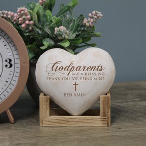 May include: White heart-shaped stone with the text "Godparents are a blessing thank you for being mine." and a small cross. The name "Benjamin" is written below the cross. The stone is sitting on a wooden stand.