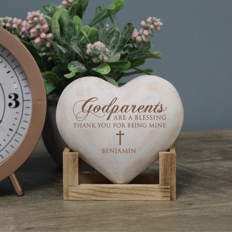 May include: White heart-shaped stone with the text "Godparents are a blessing thank you for being mine." and a small cross. The name "Benjamin" is written below the cross. The stone is sitting on a wooden stand.