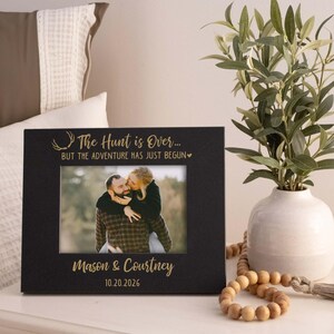 May include: A black picture frame with the text "The Hunt is Over... BUT THE ADVENTURE HAS JUST BEGUN" and the names "Mason & Courtney" with the date "10.20.2026" in gold lettering. The frame holds a photo of a couple. A white vase with greenery is to the right.