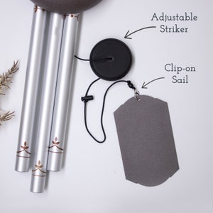 May include: Silver wind chime with brown leaf designs on the tubes. The chime has a black adjustable striker and a gray clip-on sail.