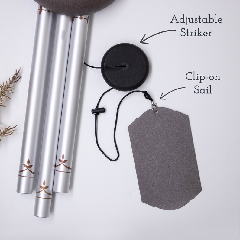 May include: Silver wind chime with brown leaf designs on the tubes. The chime has a black adjustable striker and a gray clip-on sail.