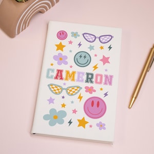 Peut inclure: Un carnet blanc avec un motif coloré représentant le nom "CAMERON" en lettres arc-en-ciel, des lunettes de soleil, des étoiles, des fleurs et des visages souriants.