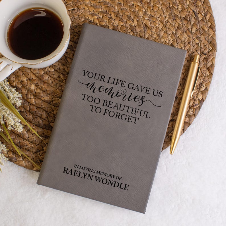 May include: A gray journal with the text "YOUR LIFE GAVE US memories TOO BEAUTIFUL TO FORGET" and "IN LOVING MEMORY OF RAELYN WONDLE". A gold pen and a cup of coffee are next to the journal. The journal is on a woven mat.