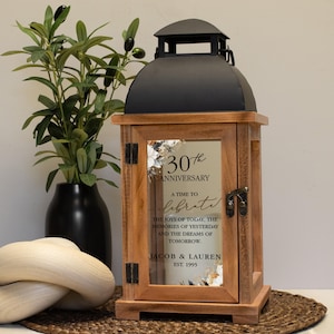 May include: A wooden lantern with a black top and a glass door. The glass door has the text "30th Anniversary, A time to celebrate" and the names "Jacob & Lauren" with the year "EST. 1995". A black vase with a plant is to the left.