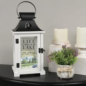 May include: White lantern with a black top and a glass window. The glass window has a watercolor painting of a lake scene and the text "Life is better at the lake. The Lowells. Lake Glendale, Pennsylvania."