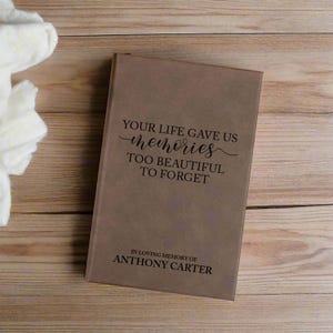 May include: A brown memorial book with the text "YOUR LIFE GAVE US memories TOO BEAUTIFUL TO FORGET" and "IN LOVING MEMORY OF ANTHONY CARTER" printed on the cover. The book is rectangular and appears to be made of leather or a similar material.