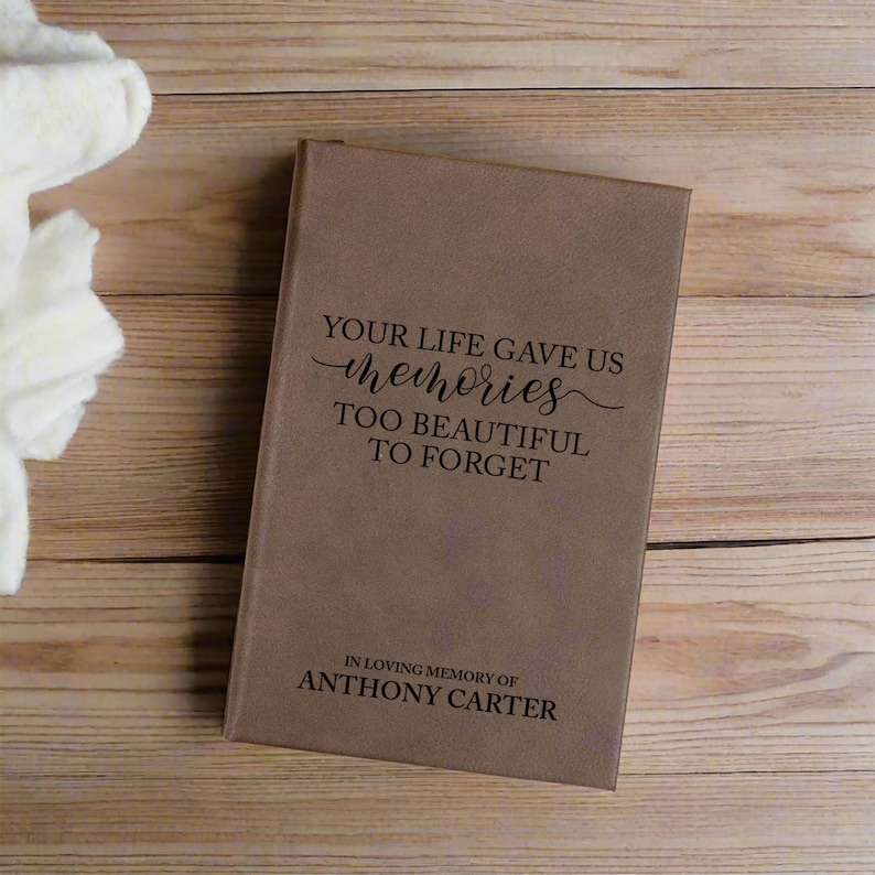 May include: A brown memorial book with the text "YOUR LIFE GAVE US memories TOO BEAUTIFUL TO FORGET" and "IN LOVING MEMORY OF ANTHONY CARTER" printed on the cover. The book is rectangular and appears to be made of leather or a similar material.